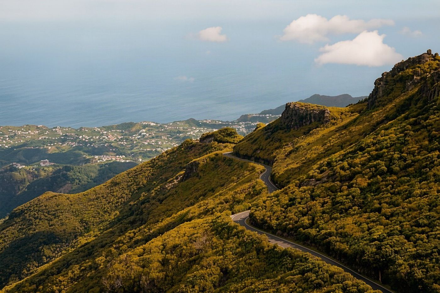 A winding asphalt road cutting through lush green mountains with clouds hovering below the peaks.